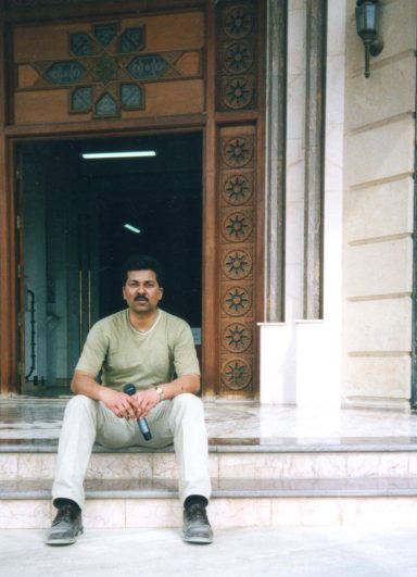 Muhammad Farhan Aslam sitting with a microphone outside the main door of Saddam Hussein’s palace in Iraq.